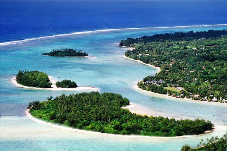 Aerial side-view image of a clear and calm Muri lagoon capturing the forest-covered islets on white sandy beaches with contrasting shades of blue waters of Rarotonga