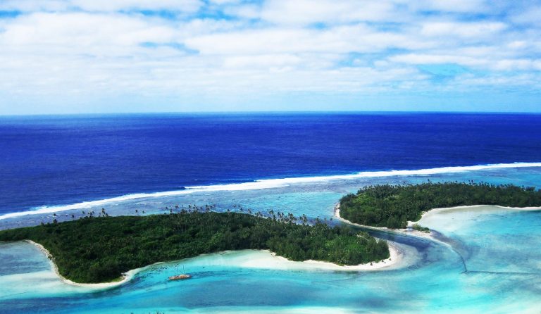 A breath-taking aerial view the forest-covered islets in Muri on a glowing white sandy beach with contrasting all-shades-of-blue waters of Rarotonga, also capturing a sailing boat berthed offshore