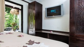 image of a flat TV screen mounted on to a wall between two tall carved-Polynesian-style deep brown colour wardrobes, a view from the super king bed