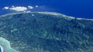 Aerial view of Rarotonga Island featuring a clear lagoon surrounded by coral reefs that separates the island from the appealing exquisite shade of rich royal blue of the ocean, evoking its depth