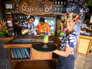 waitress happily demonstrating her skills holding a serving tray with a glass of cocktail, also capturing a busy and cheerful bartenders in the background