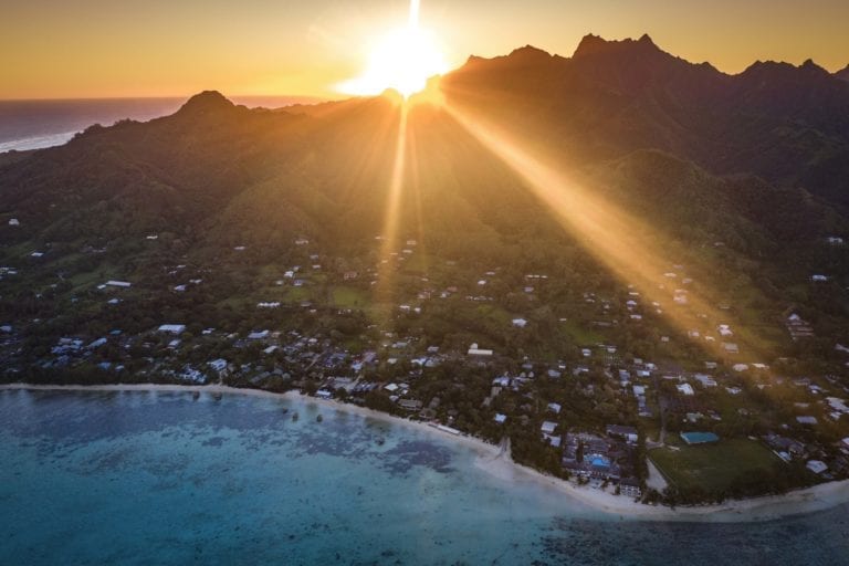 A stunning aerial image of the South-Eastern side of Rarotonga Island capturing a glorious and breathtaking sunset in the background which illuminates the horizon