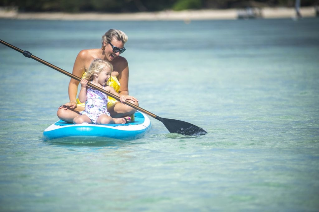 Image of a toddler enjoy paddling on a paddleboard with the company of her mom coaching the moves