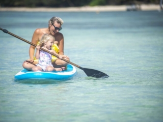 Image of a toddler enjoy paddling on a paddleboard with the company of her mom coaching the moves