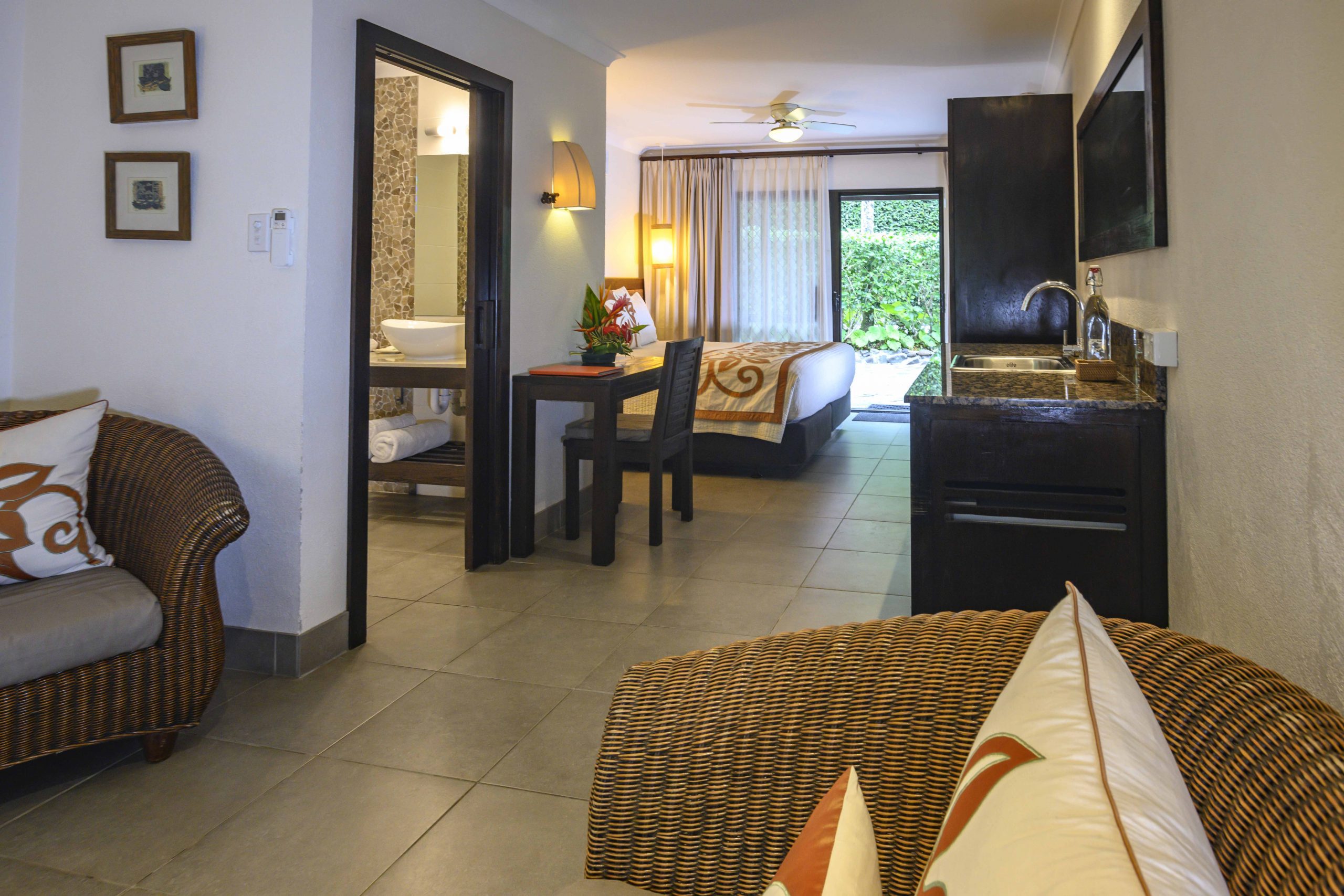 image of a luminous Premium Garden Suite taken from the lounge area featuring a super king bed, a desk and sink also capturing a partial view of the tropical garden in the front yard